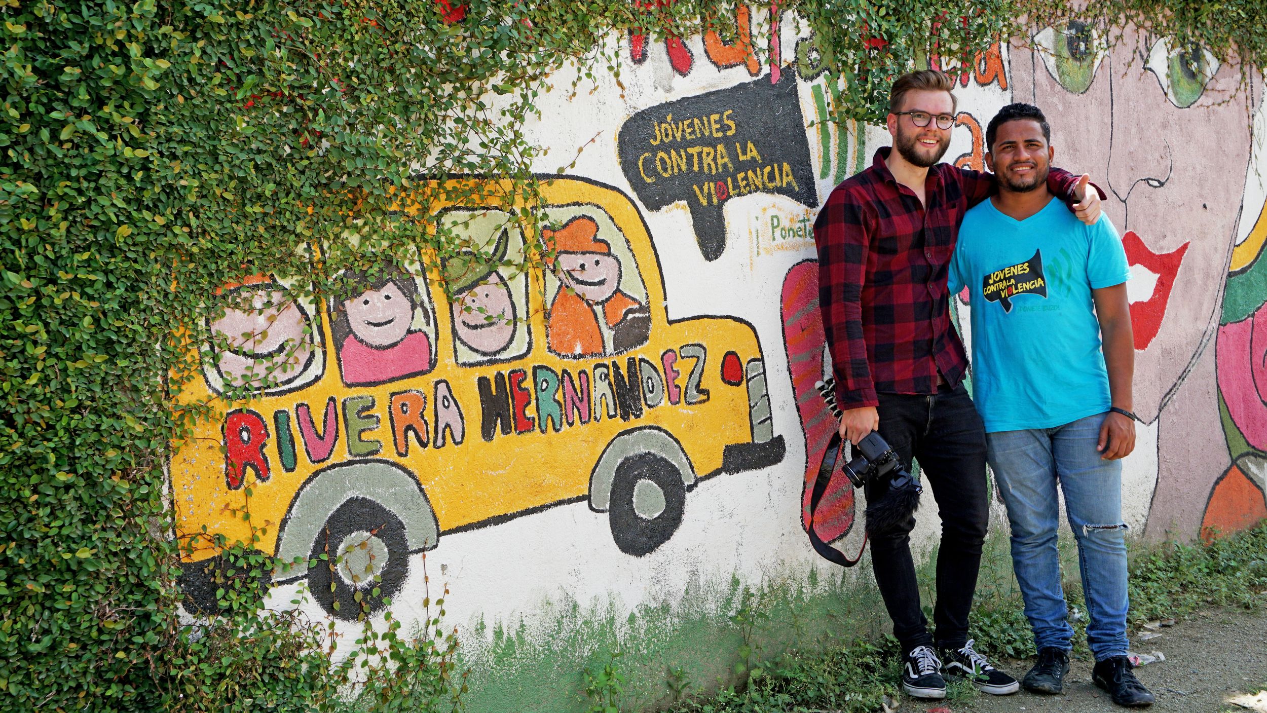 Flo steht mit einem Mann aus Honduras vor einer Wand mit einem aufgemalten Auto und spanischen Wörtern