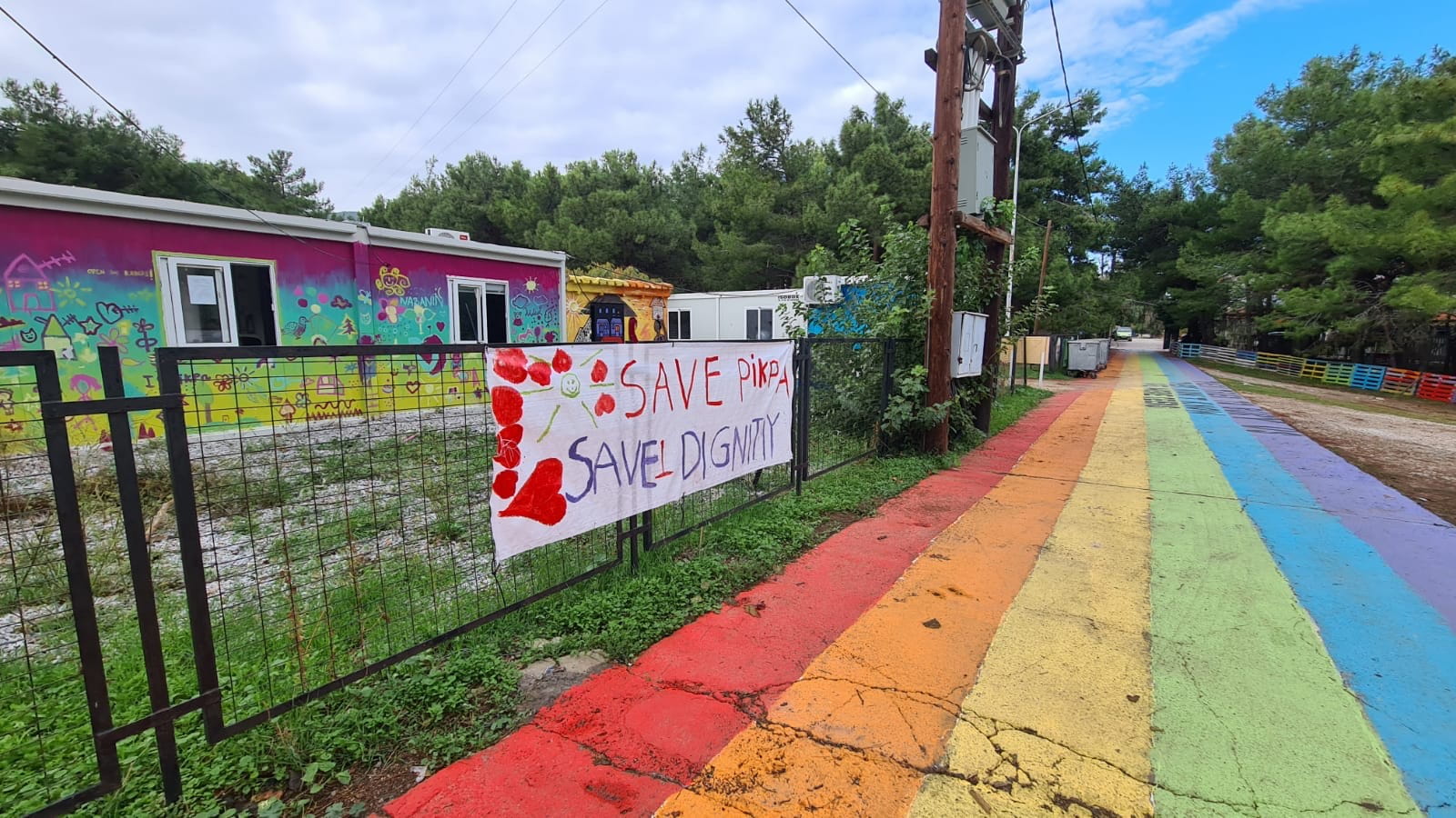 Eine bunte Straße in Regenbogenfarben führt an einem Zaun entlang, auf einem Banner steht: Rette Pikpa, rette Würde (Quelle: LESOL) Eine bunte Straße in Regenbogenfarben führt an einem Zaun entlang, auf einem Banner steht: Rette Pikpa, rette Würde (Quelle: LESOL)