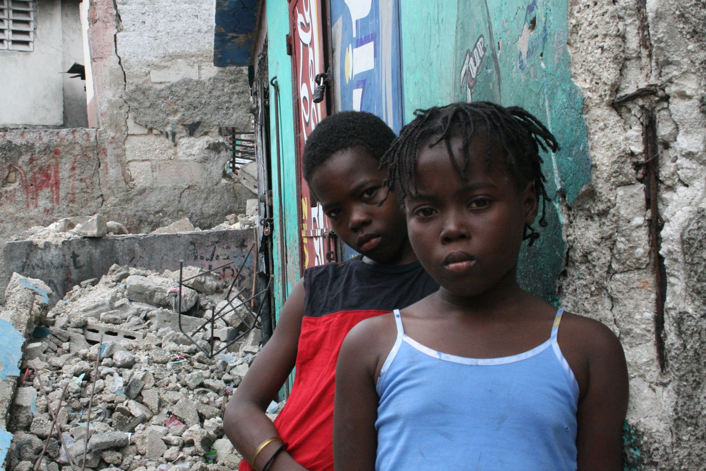 Ein Mädchen und ein Junge aus Haiti lehnen ernst dreinschauend an einer Wand vor Trümmern. (Quelle: Jürgen Schübelin)
