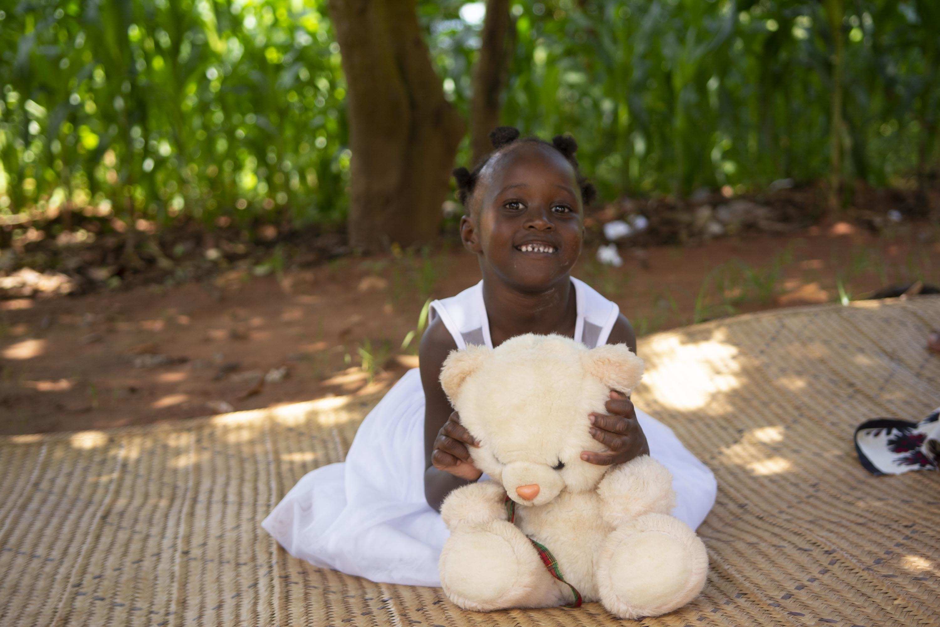 Malawi: Ein Mädchen sitzt auf einer Strohmatte und hält lächelnd einen Teddybär im Arm. (Quelle: Christian Nusch)