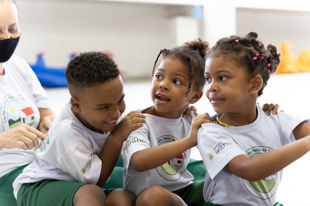 Drei Kinder sitzen lachend hintereinander und fassen sich an den Schultern. (Quelle: Christian Nusch)  Drei Kinder sitzen lachend hintereinander und fassen sich an den Schultern. (Quelle: Christian Nusch)