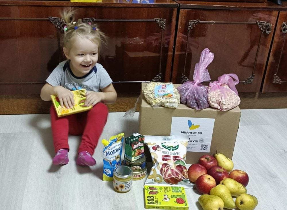 Ein kleines Mädchen sitzt lachend neben einem Lebensmittelpaket. (Quelle: Myrne Nebo)