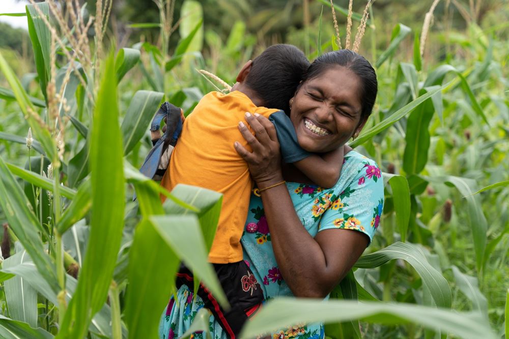 Eine Mutter aus einer Frauengruppe steht in ihrem Maisfeld und umarmt lachend ihren kleinen Sohn (Quelle: Christian Nusch)