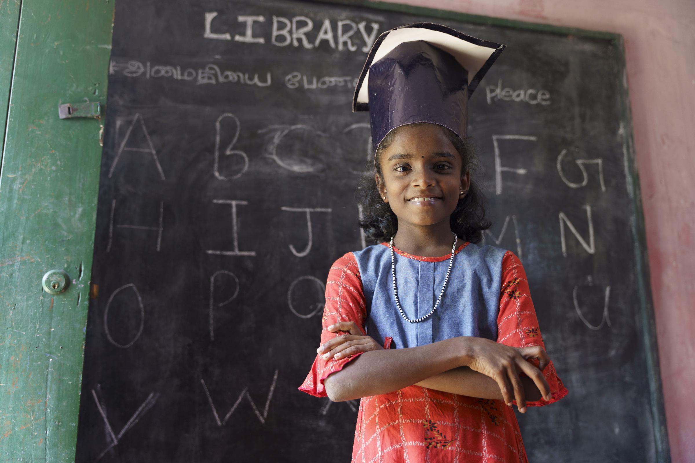 Ein Mädchen aus Sri Lanka steht in der Schule vor einer Tafel und lacht in die Kamera (Quelle: Christian Nusch)