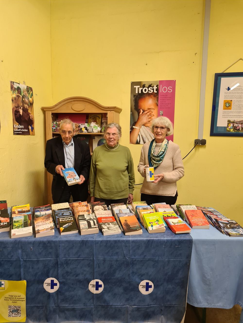 Ein Büchertisch, dahinter 3 Mitglieder des Arbeitskreises, Kindernothilfe-Poster an der Wand (Quelle: Christopher Vent) Ein Büchertisch, dahinter 3 Mitglieder des Arbeitskreises, Kindernothilfe-Poster an der Wand (Quelle: Christopher Vent)