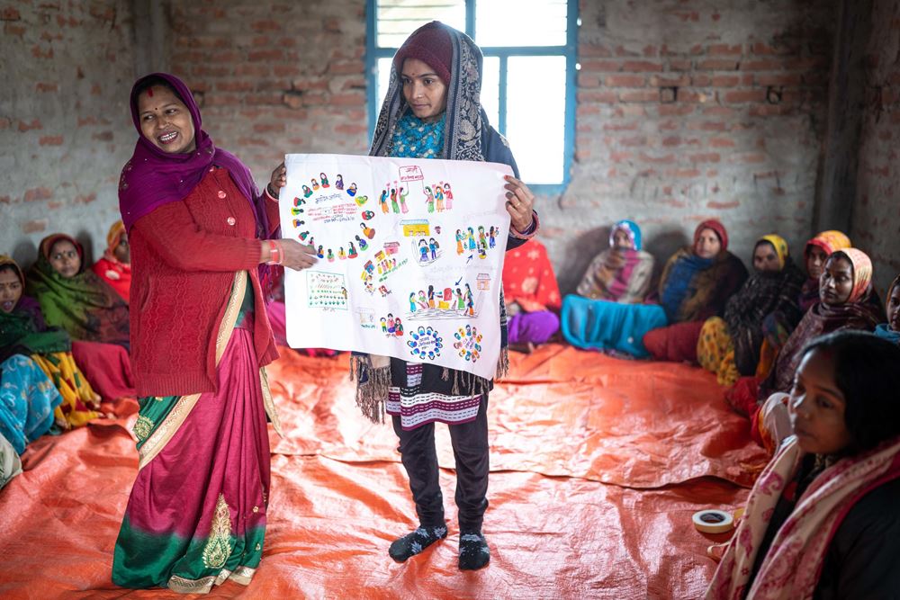 Eine Frauengruppe sitzt an der Wand entlang auf einer orangen Plane, in der Mitte stehen zwei Frauen mit einem selbst gemalten Schaubild (Quelle: Jakob Studnar) Eine Frauengruppe sitzt an der Wand entlang auf einer orangen Plane, in der Mitte stehen zwei Frauen mit einem selbst gemalten Schaubild (Quelle: Jakob Studnar)