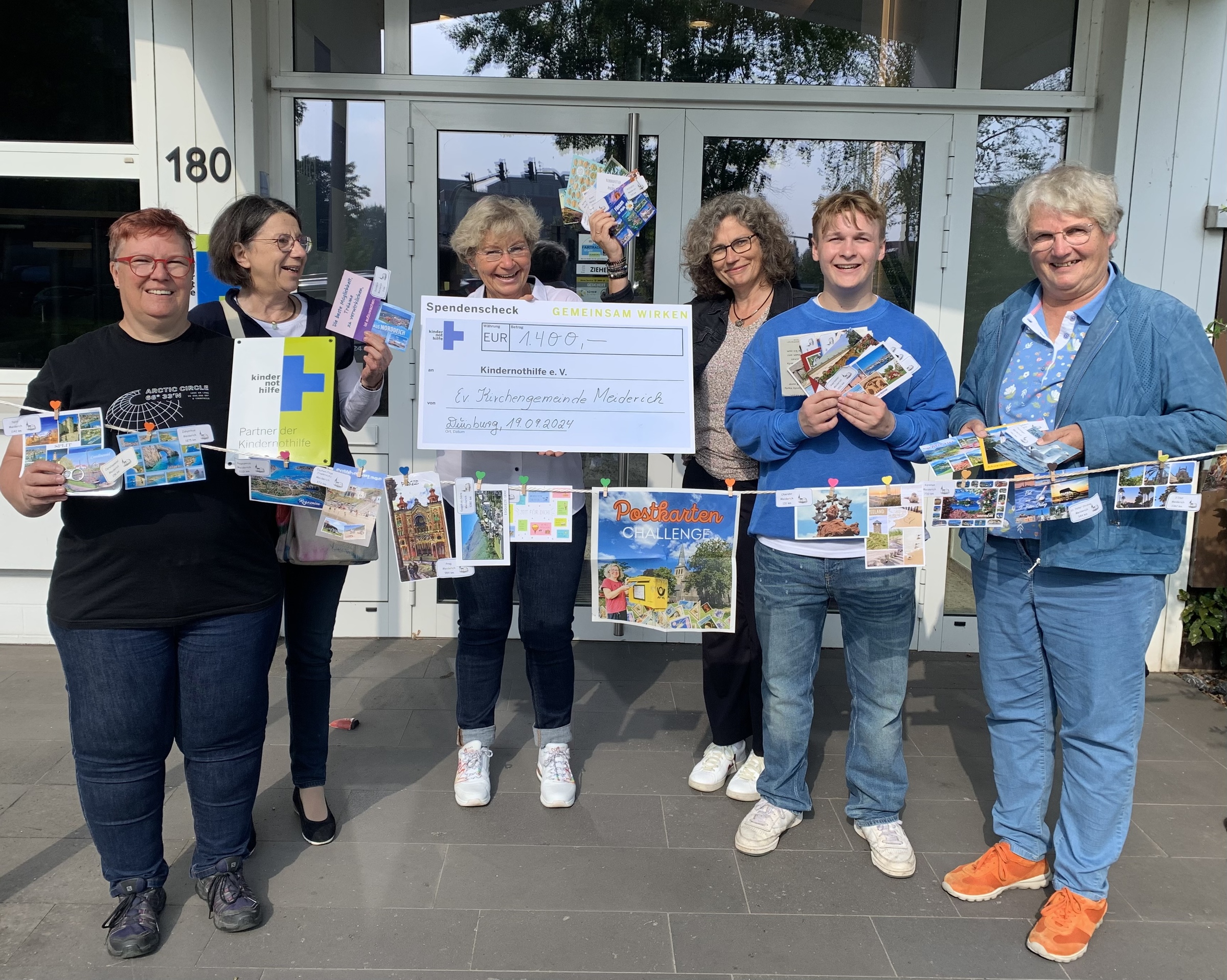 Sechs Personen stehen mit einer Leine, an der Postkarten hängen, und einem großen Scheck vor dem Eingang der Kindernothilfe (Quelle: Sophie Rutter) Sechs Personen stehen mit einer Leine, an der Postkarten hängen, und einem großen Scheck vor dem Eingang der Kindernothilfe (Quelle: Sophie Rutter)