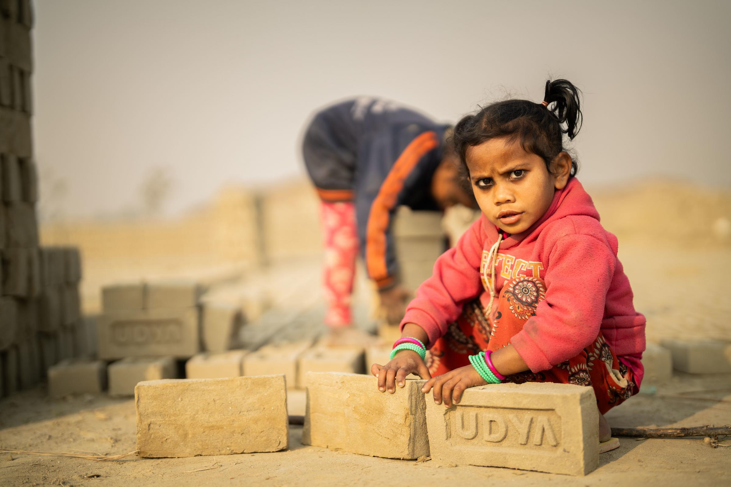 Ein kleines Mädchen schaut in die Kamera. Sie hockt auf dem Boden und hält zwei große Ziegelsteine in der Hand. Foto : Jakob Studnar / Kindernothilfe.
