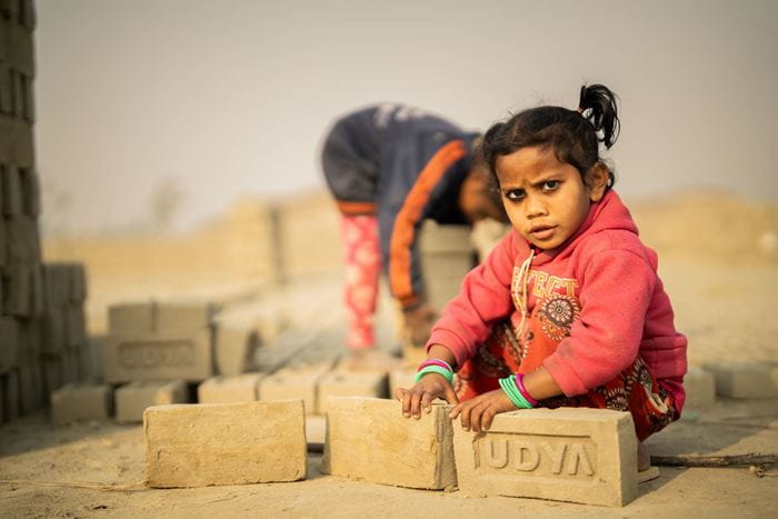 Ein kleines Mädchen schaut in die Kamera. Sie hockt auf dem Boden und hält zwei große Ziegelsteine in der Hand. Foto : Jakob Studnar / Kindernothilfe. Ein kleines Mädchen schaut in die Kamera. Sie hockt auf dem Boden und hält zwei große Ziegelsteine in der Hand. Foto : Jakob Studnar / Kindernothilfe.