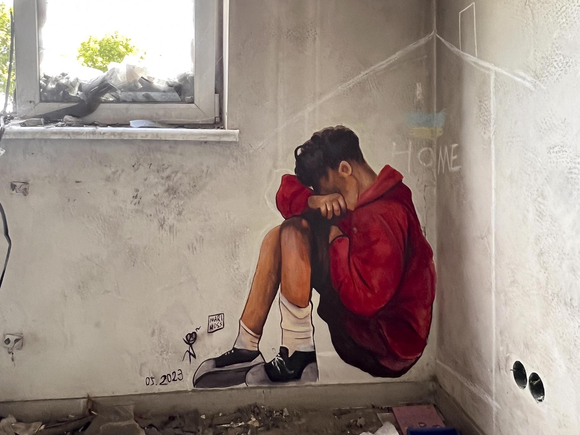 Auf einer Wand in einem zerstörten Haus ist ein Junge gemalt, der auf dem Boden sitzt. Er hat seinen Kopf auf seine Arme gelegt und sich auf seinem Knie abgestützt. Foto: Stanislav Shulimov Auf einer Wand in einem zerstörten Haus ist ein Junge gemalt, der auf dem Boden sitzt. Er hat seinen Kopf auf seine Arme gelegt und sich auf seinem Knie abgestützt. Foto: Stanislav Shulimov