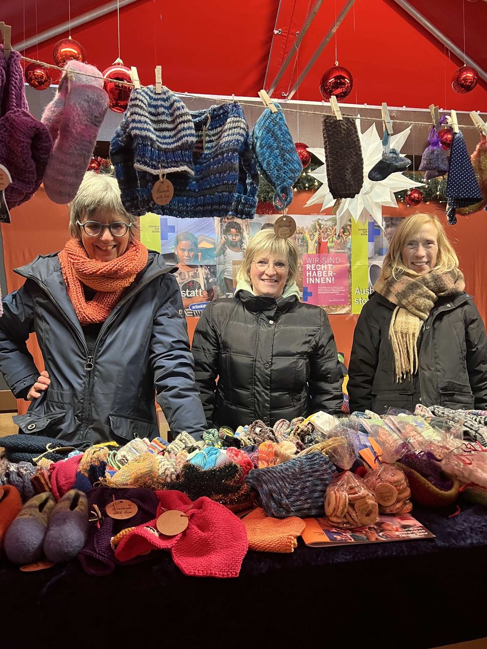 Drei Frauen in einer Weihnachtsmarkt-Bude, vor ihnen liegen und hängen ganz viele Stricksachen (Quelle: privat) Drei Frauen in einer Weihnachtsmarkt-Bude, vor ihnen liegen und hängen ganz viele Stricksachen (Quelle: privat)