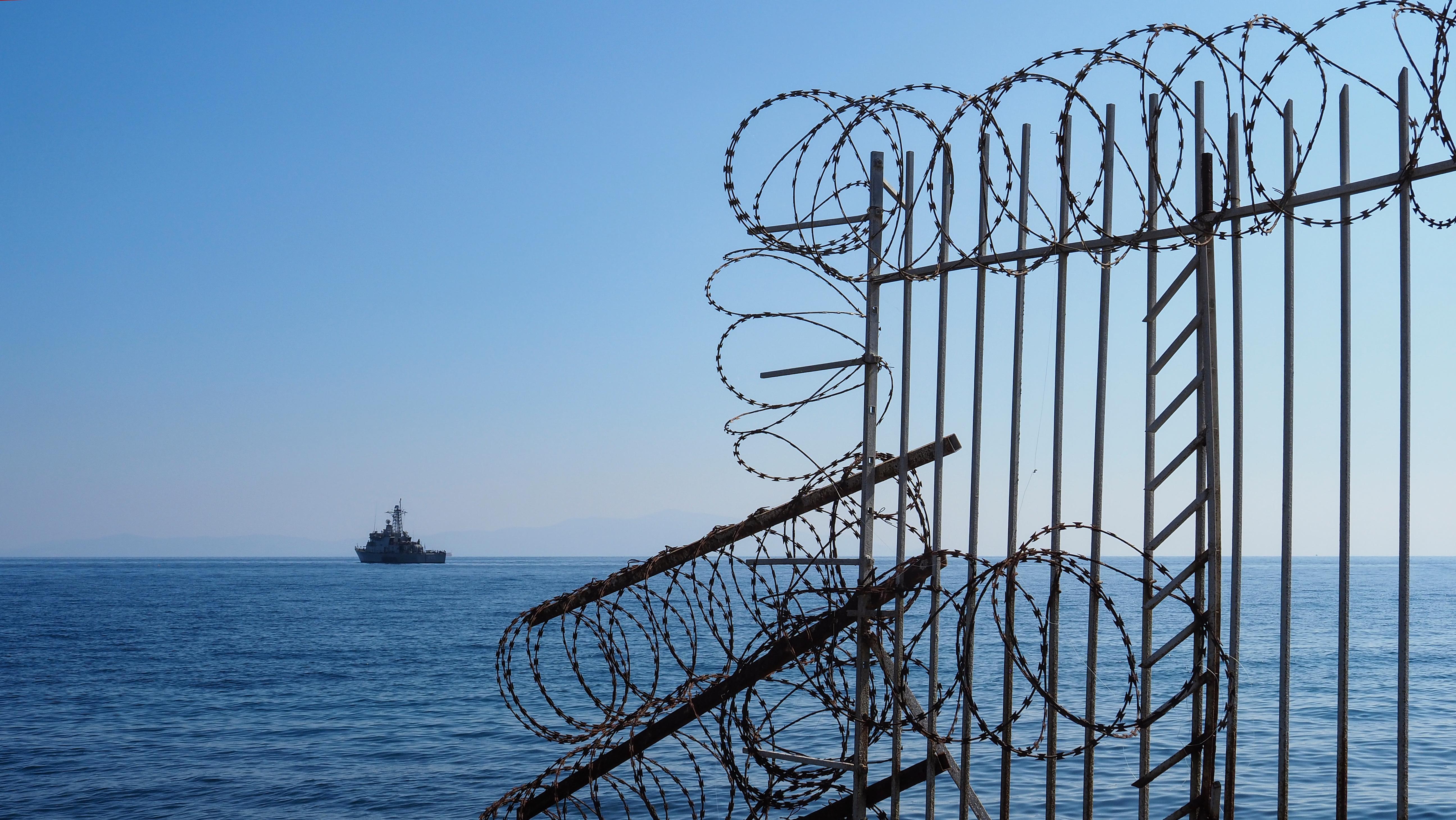 Symbolbild Grenzzaun: Blick auf das Ägäische Meer und ein Kriegsschiff, unmittelbar neben dem mit Stacheldraht umzäunten Lager Mavrovouni aufgenommen (Quelle: Knut Bry) Symbolbild Grenzzaun: Blick auf das Ägäische Meer und ein Kriegsschiff, unmittelbar neben dem mit Stacheldraht umzäunten Lager Mavrovouni aufgenommen (Quelle: Knut Bry)