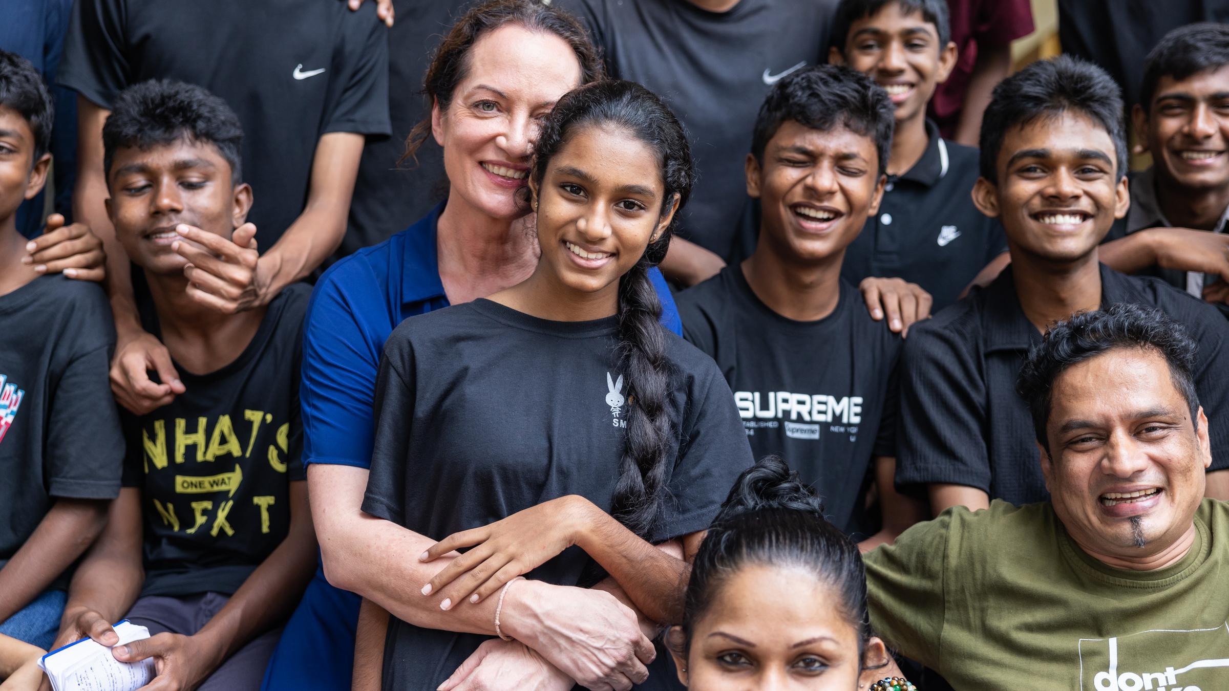 Schauspielerin und Kindernothilfe-Botschafterin Natalia Wörner umarmt ein Mädchen. Um sie herum sitzen weitere Jugendliche und Erwachsene. Alle schauen in die Kamera und lächeln. Foto: Sharni Jayawardena