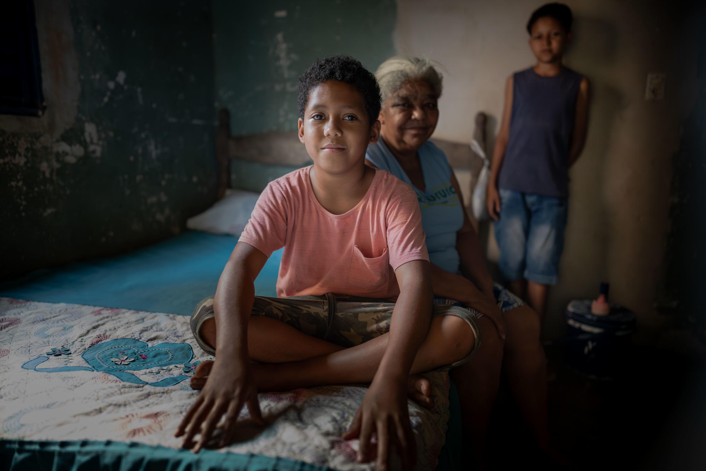 Ricardo sitzt auf seinem Bett. Dahinter sitzt seine Oma, sein Bruder steht weiter hinten. Alle schauen in die Kamera. Foto: Jakob Studnar