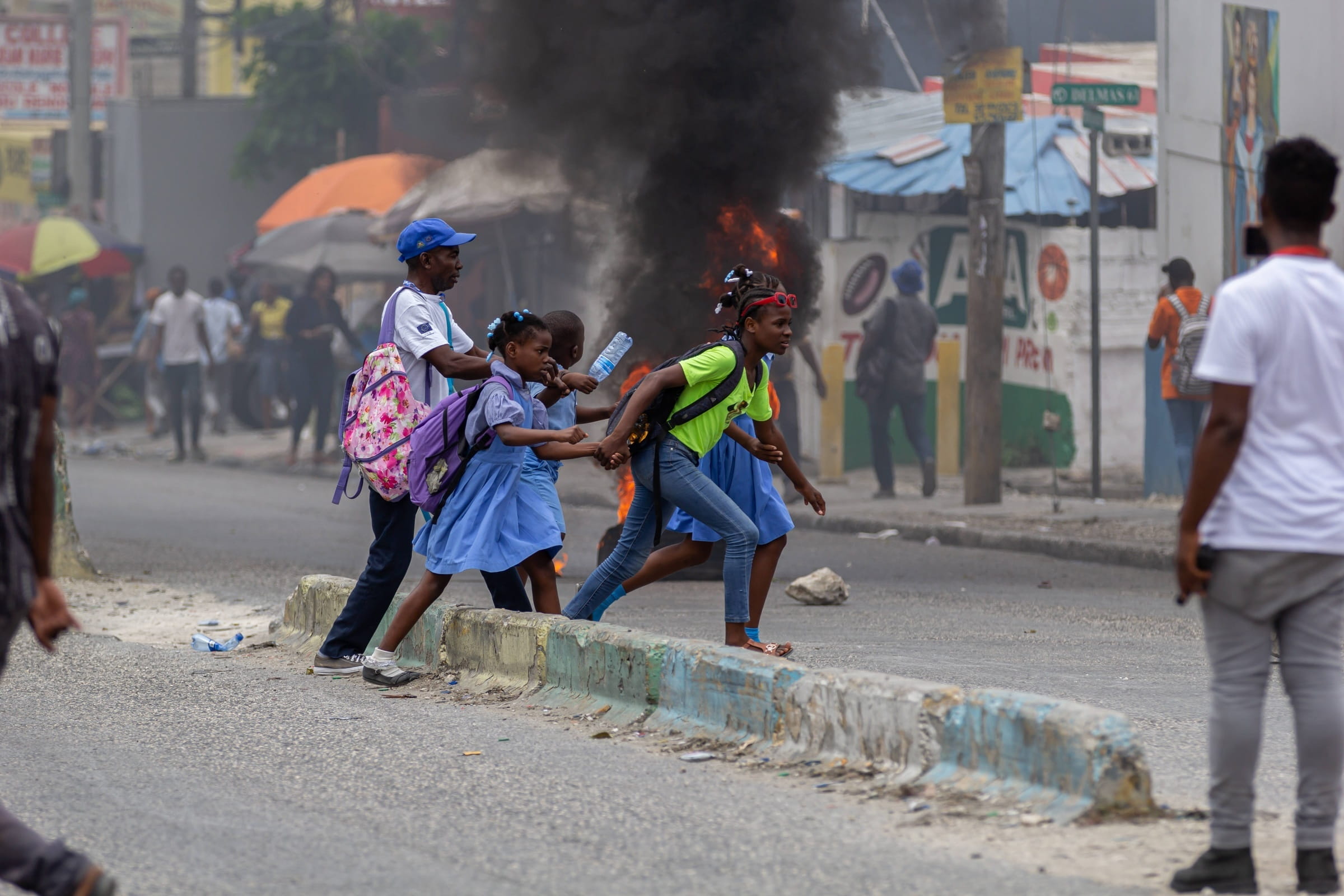 Kinder eilen über eine Straße in Port-Au-Prince, Haiti. Im Hintergrund eine hohe Flamme mit einer starken schwarzen Rauchwolke. Offene Gewalt auf den Straßen. (Quelle: IMAGO / Anadolu Agency)