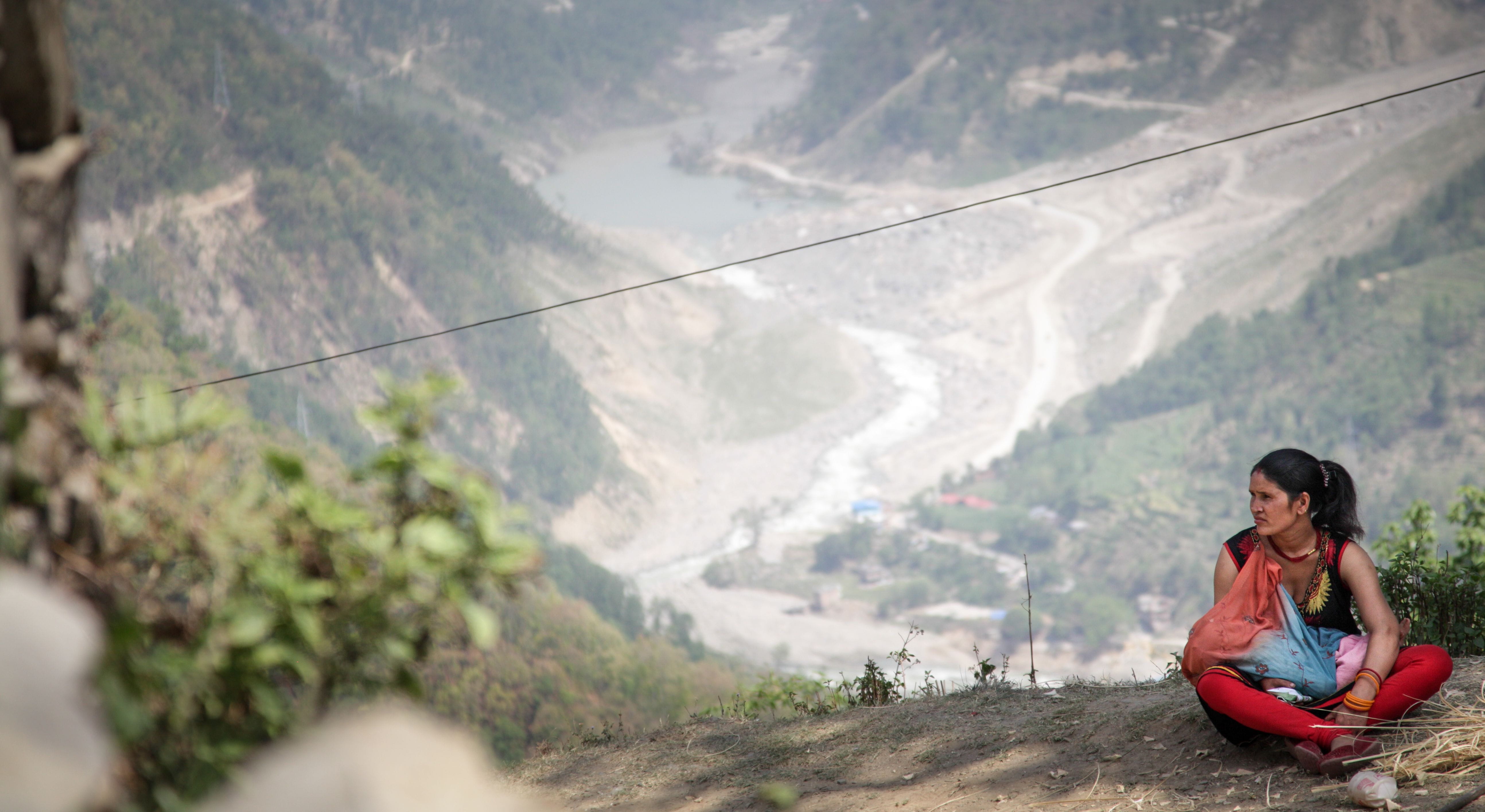 Symbolfoto Nepal: Eine Frau sitzt auf dem Boden auf einem Berg. Im Hintergrund ist ein Fluss in einem Tal (Quelle: Bastian Strauch)