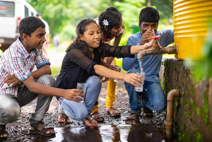 Vier Jugendliche aus Indien füllen lachend sauberes Wasser aus einem Brunnen in Gläser. (Quelle: Jakob Studnar)