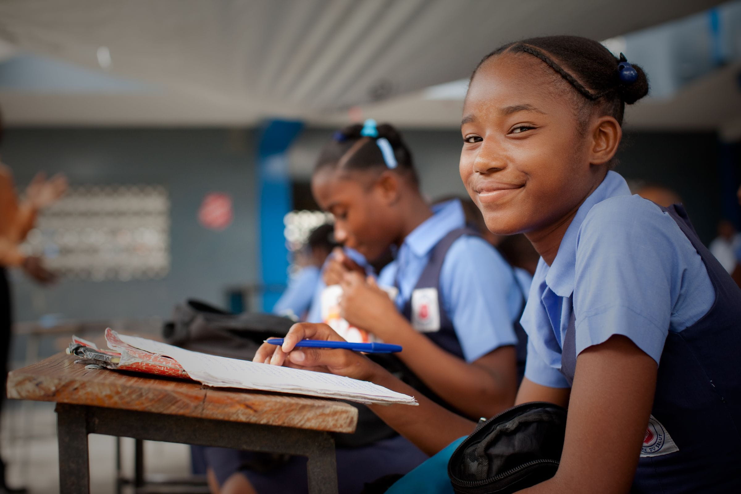 Ein Mädchen aus Haiti sitzt in eine blaue Schuluniform gekleidet in einem Klassenzimmer und lächelt. (Quelle: Jakob Studnar)