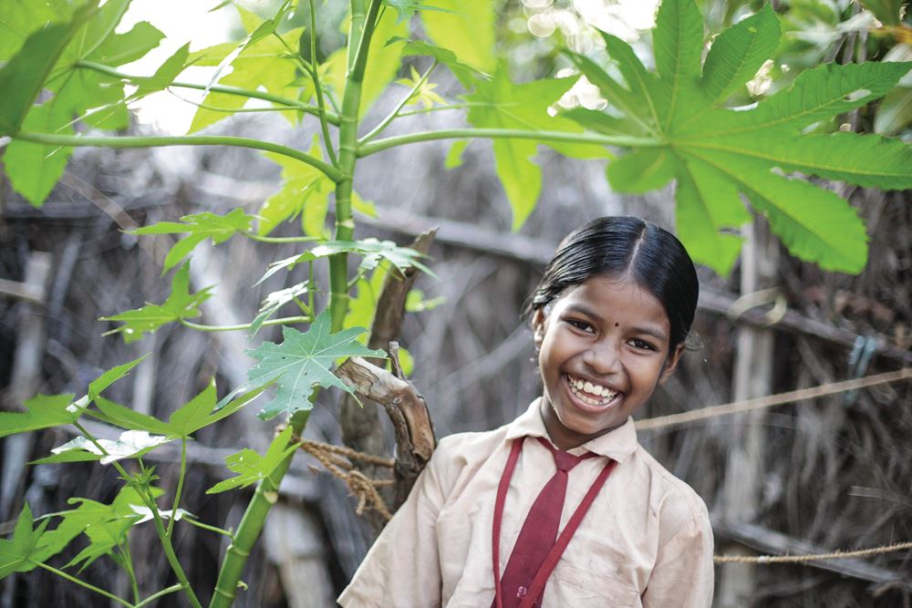 Ein indisches Mädchen steht unter einer Pflanze und lacht in die Kamera. (Quelle: Bastian Strauch)