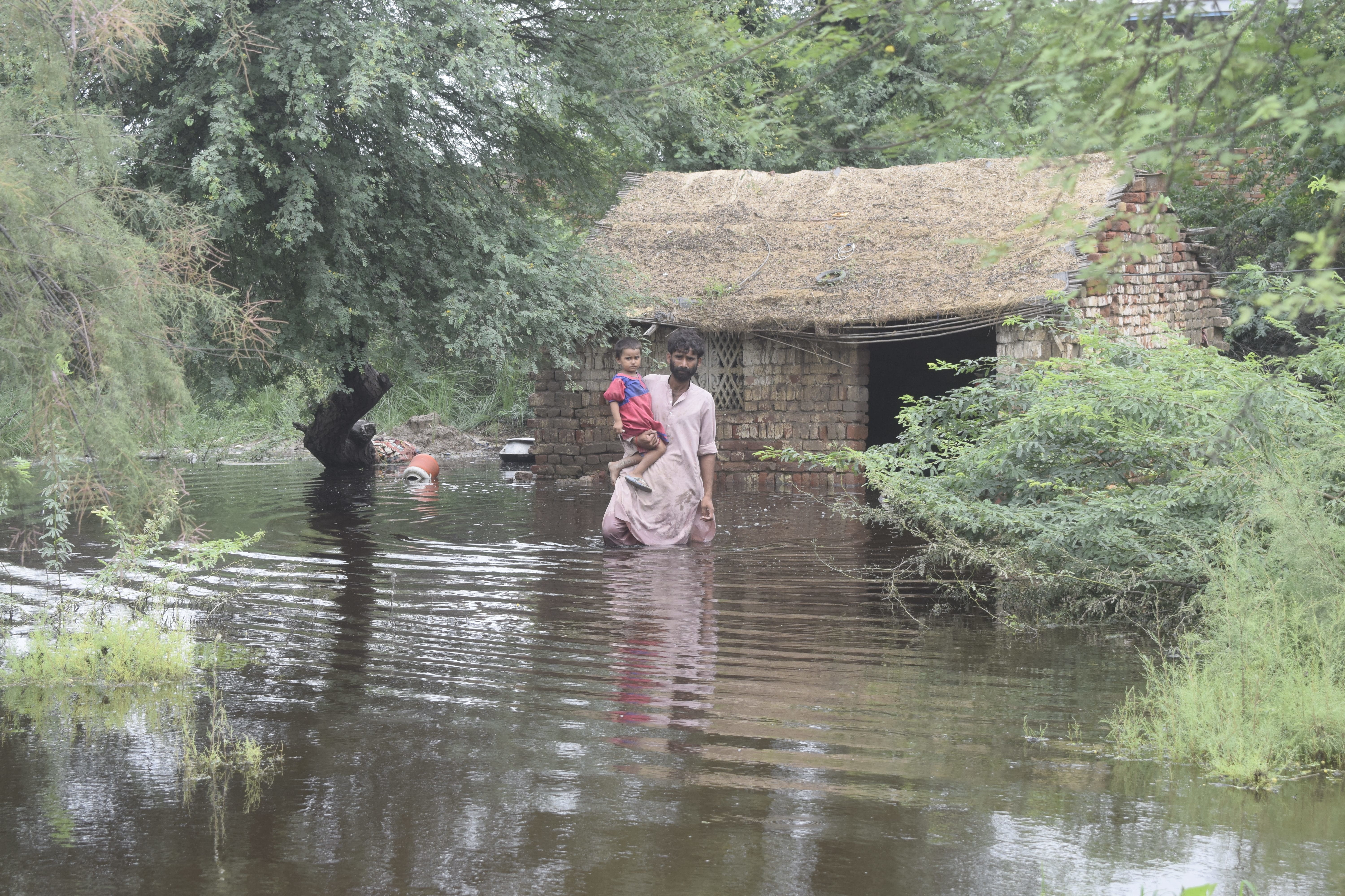 Symbolfoto: Flut, Ein Mann mit einem Kind auf dem Arm steht in hüfthohem Wasser vor einem überschwemmten Haus (Quelle: Kindernothilfe-Partner)