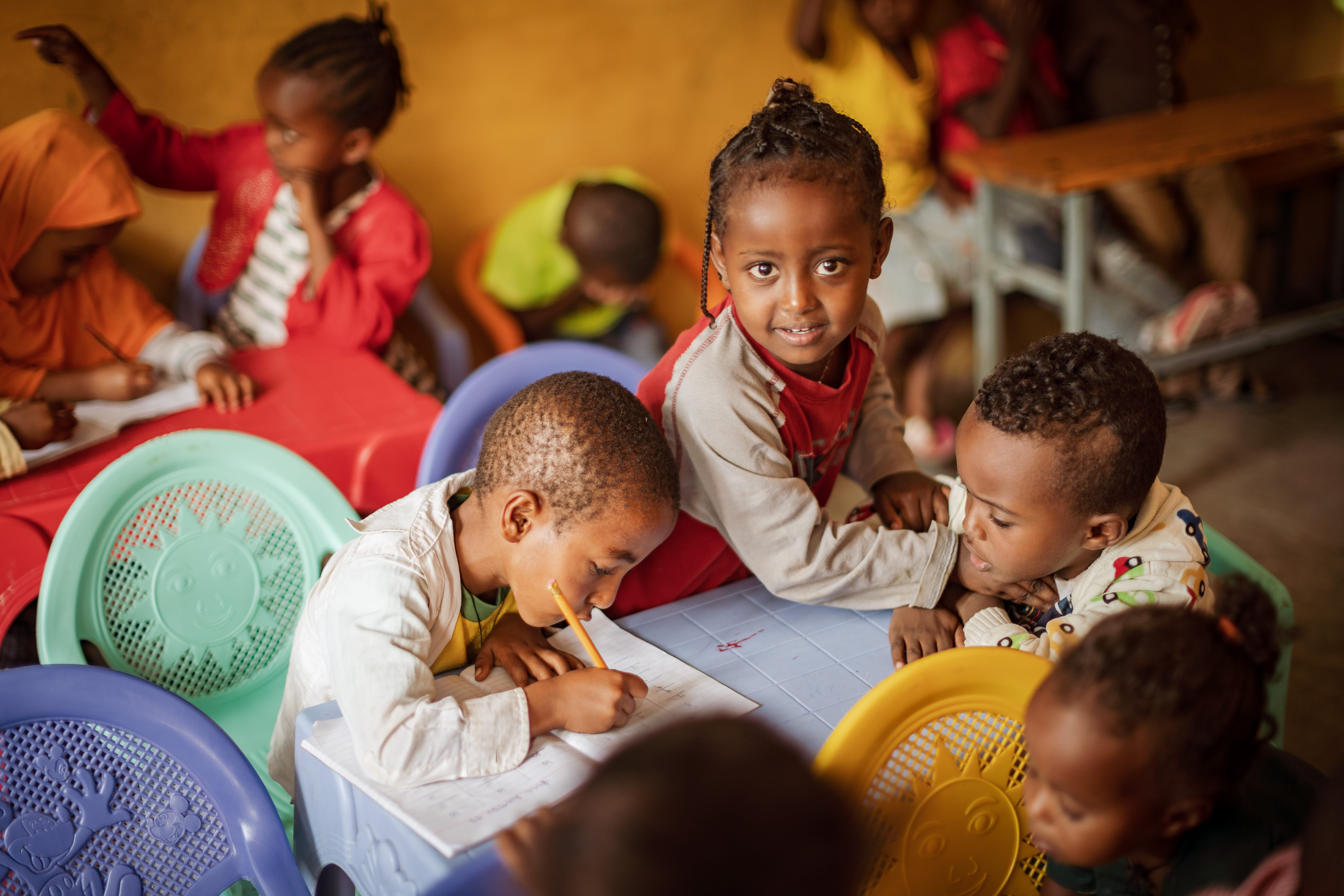 Ein paar Kinder sitzen um ein Schulpult, eins schreibt in ein Heft, eins sieht zu, eins blickt die Betrachterin fröhlich und neugierig an  (Bildquelle: Jakob Studnar)