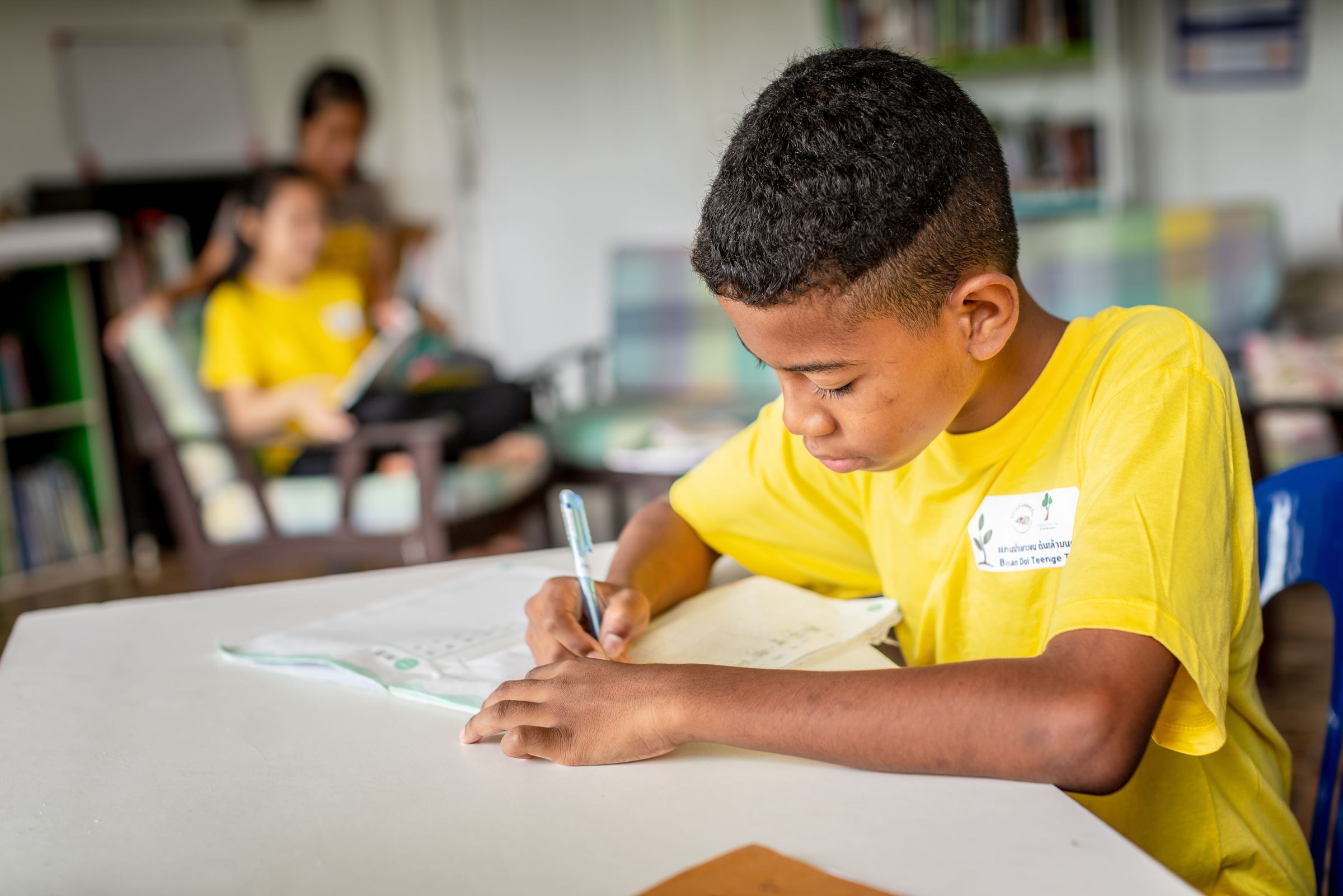 Ein Junge in gelbem T-Shirt beugt sich über ein Heft und schreibt. (Quelle: Jakob Studnar)