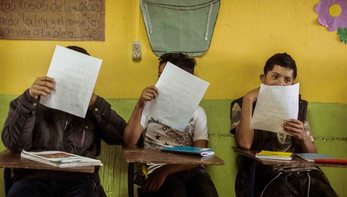 Drei Kinder in einem Klassenraum mit einer gelben Wand sitzen auf Stühlen mit Pult und halten ein Blatt Papier hoch, das sie lesen  (Bildquelle: Jakob Studnar)