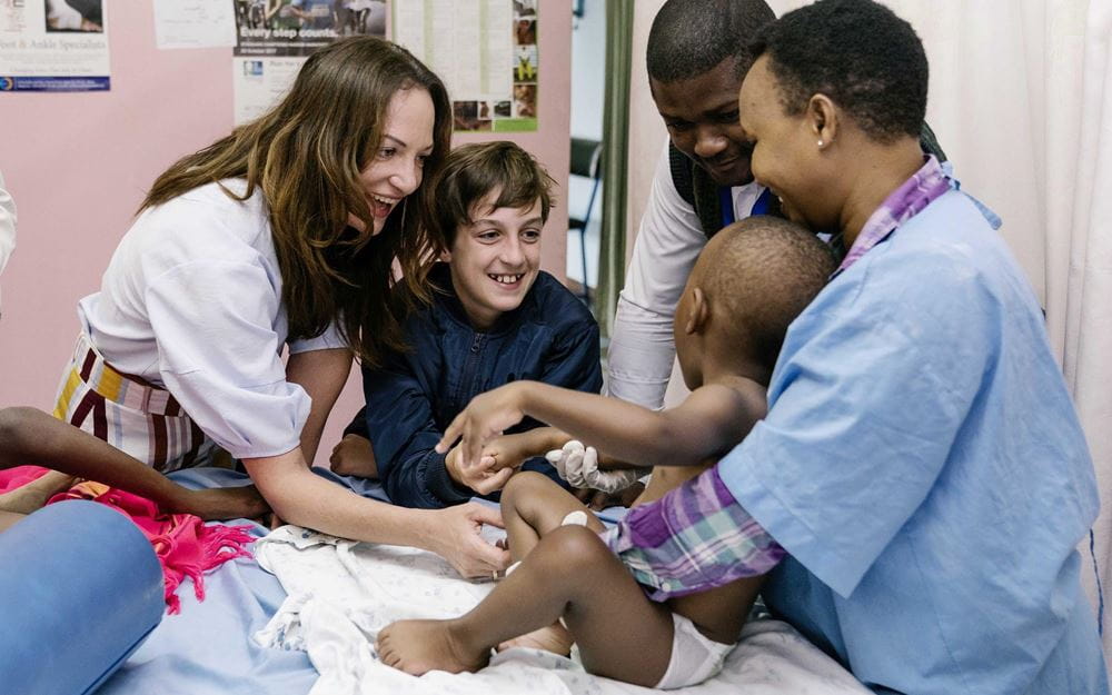 Reportage Kenia: Ein Alltag auf neun Quadratmetern; Foto: Natalia Woerner und Jacob lächeln Kind an, das von Frau mit Kittel gehalten wird (Quelle: Dan Zoubek / Kindernothilfe)  Reportage Kenia: Ein Alltag auf neun Quadratmetern; Foto: Natalia Woerner und Jacob lächeln Kind an, das von Frau mit Kittel gehalten wird (Quelle: Dan Zoubek / Kindernothilfe)