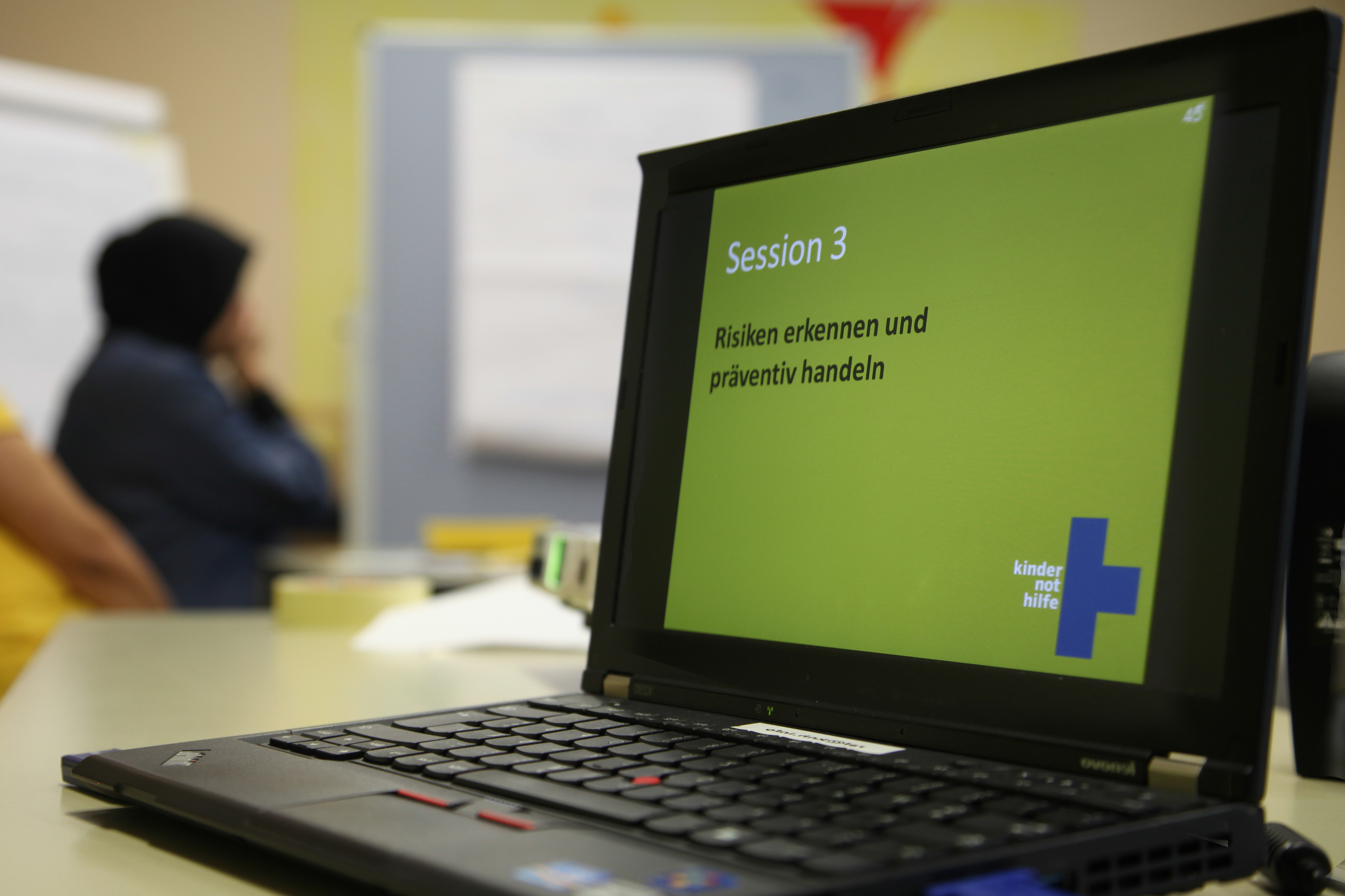 Laptop mit geöffneter Powerpoint-Folie – im Hintergrund Schulungsteilnehmerin (Quelle: Ludwig Grunewald, Kindernothilfe).