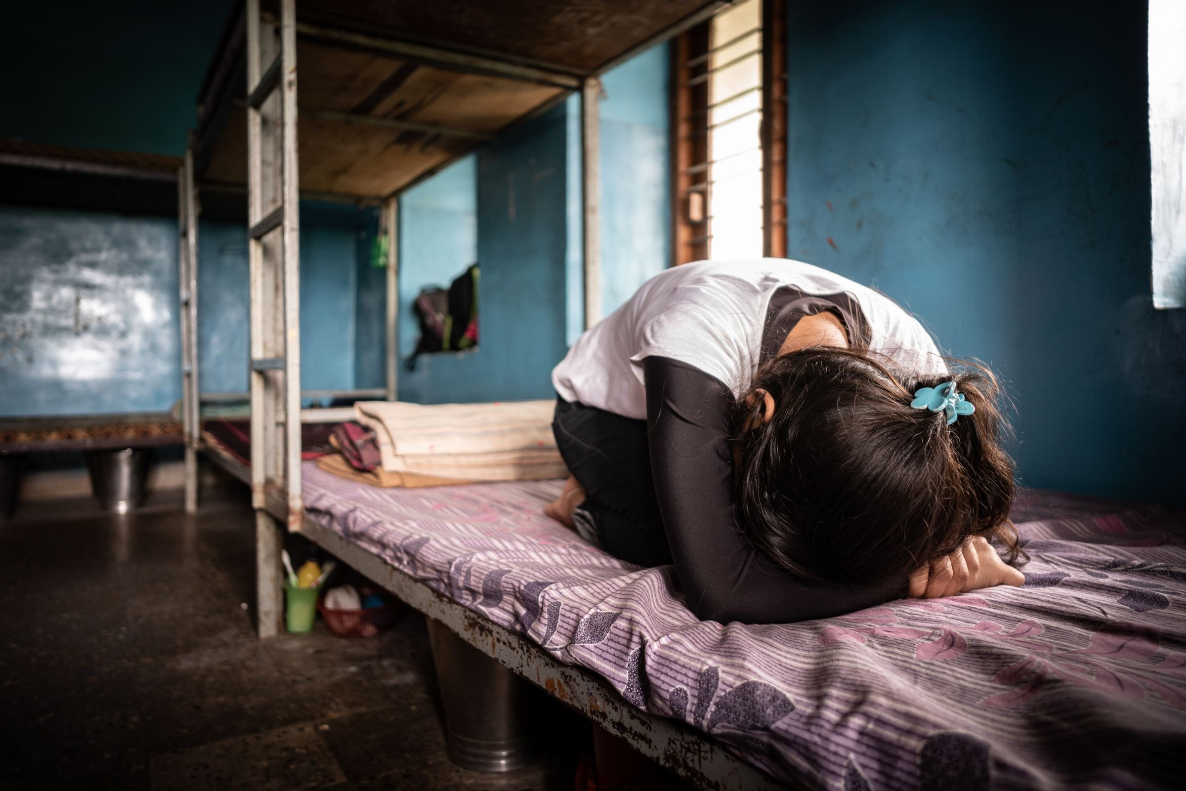 Ein Mädchen liegt zusammengekauert auf einem einfachen Bett und verbirgt ihr Gesicht in ihren Armen. (Quelle: Jakob Studnar)