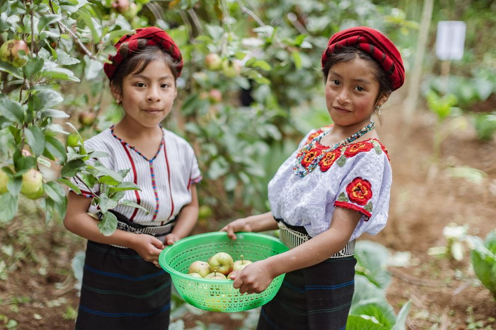 Zwei Mädchen in traditioneller Kleidung aus Guatemala halten einen Apfelkorb im Arm. (Quelle: Jakob Studnar)