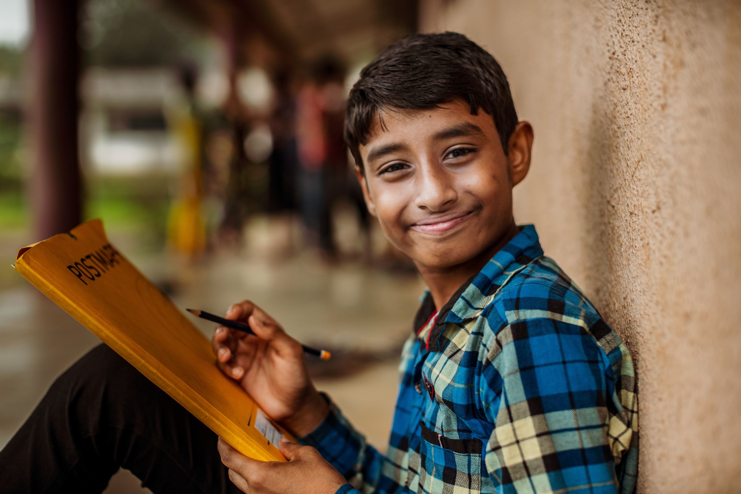 ein Junge mit einem Heft und einem Bleistift in der Hand lehnt an einer Hauswand und lächelt breit. (Bildquelle: Jakob Studnar)