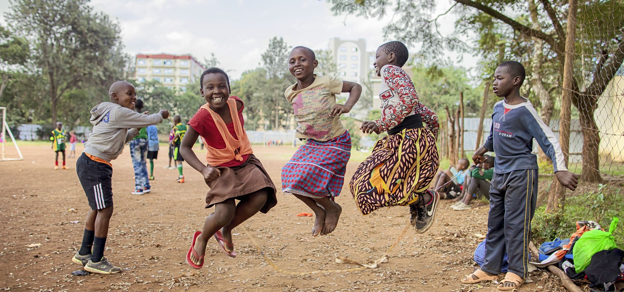 Kinder in Kenia springen auf einem Sandplatz wild durcheinander, im Hintergrund Bäume und große Häuser (Bildquelle: Lars Heidrich)